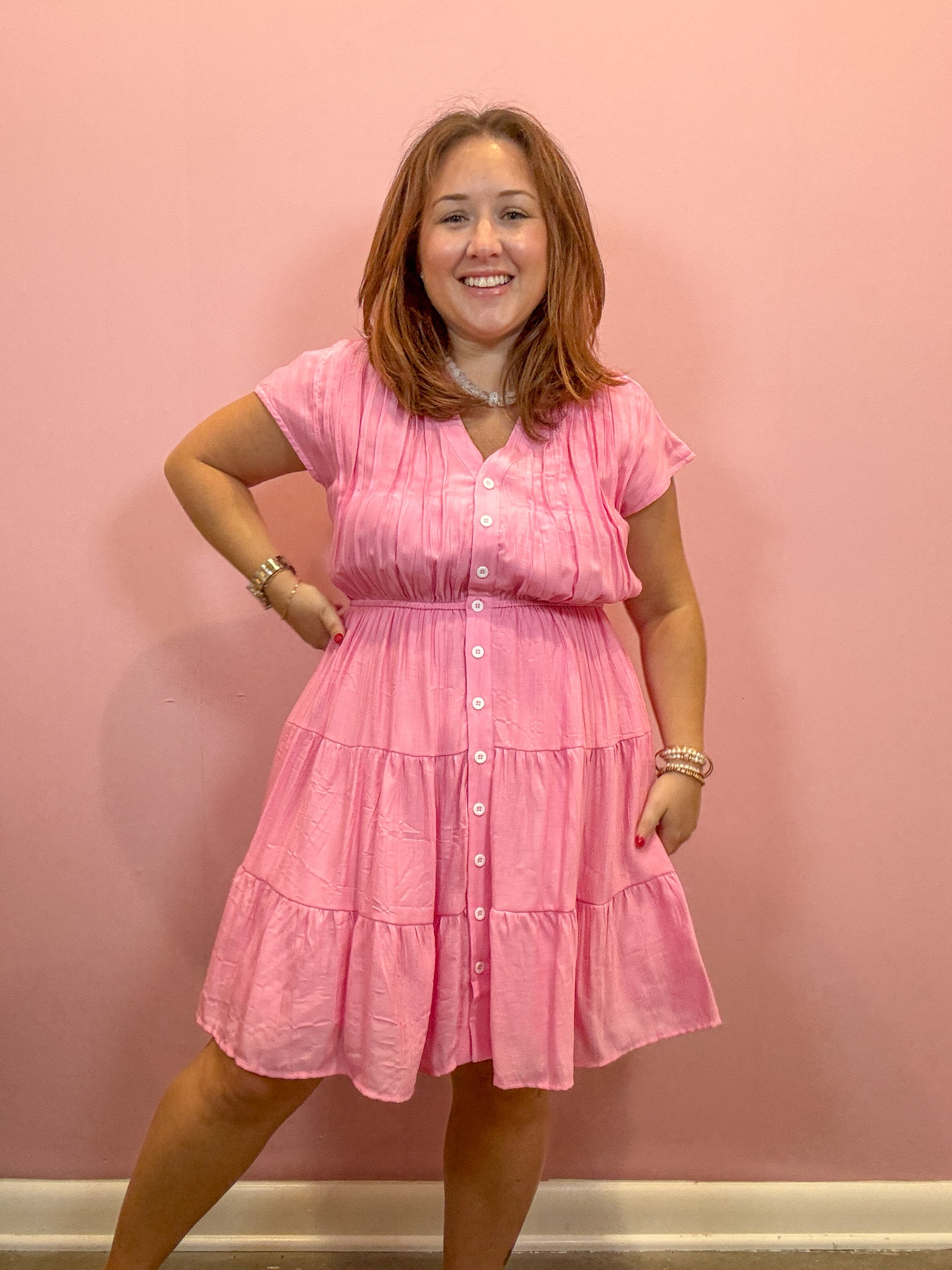 A woman modeling a pink tiered short-sleeve button-up dress against a solid pink wall.