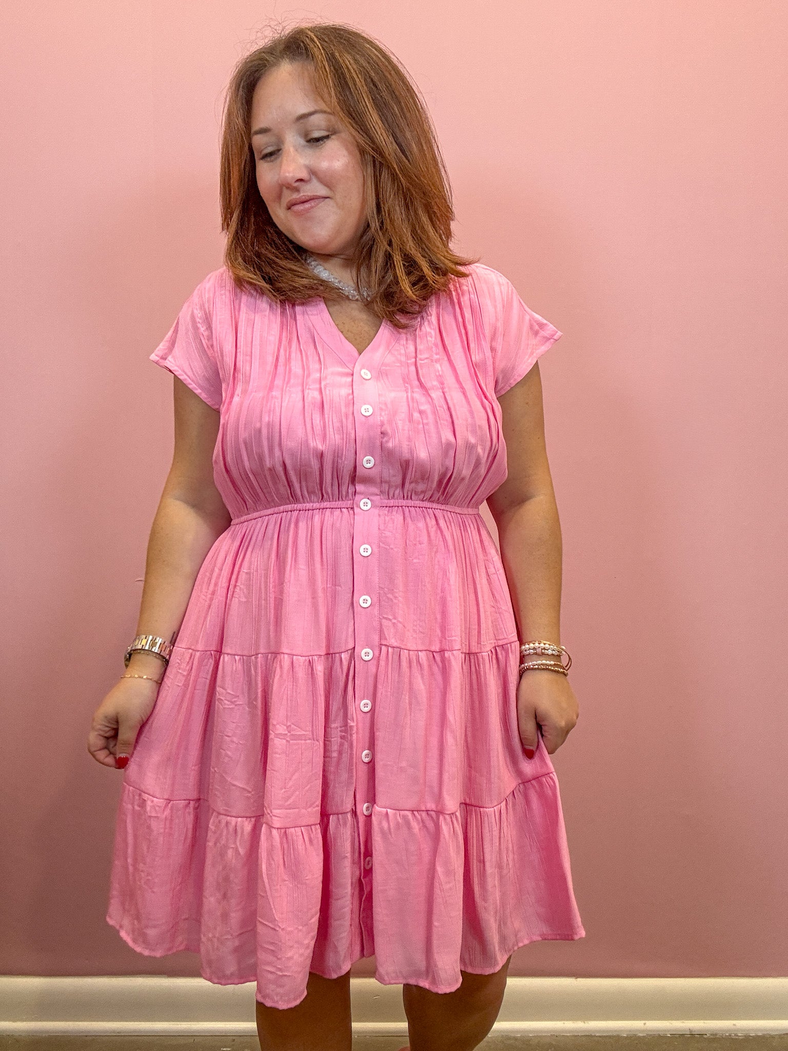 A woman wearing a pink tiered short-sleeve button-front dress, standing against a solid pink wall.