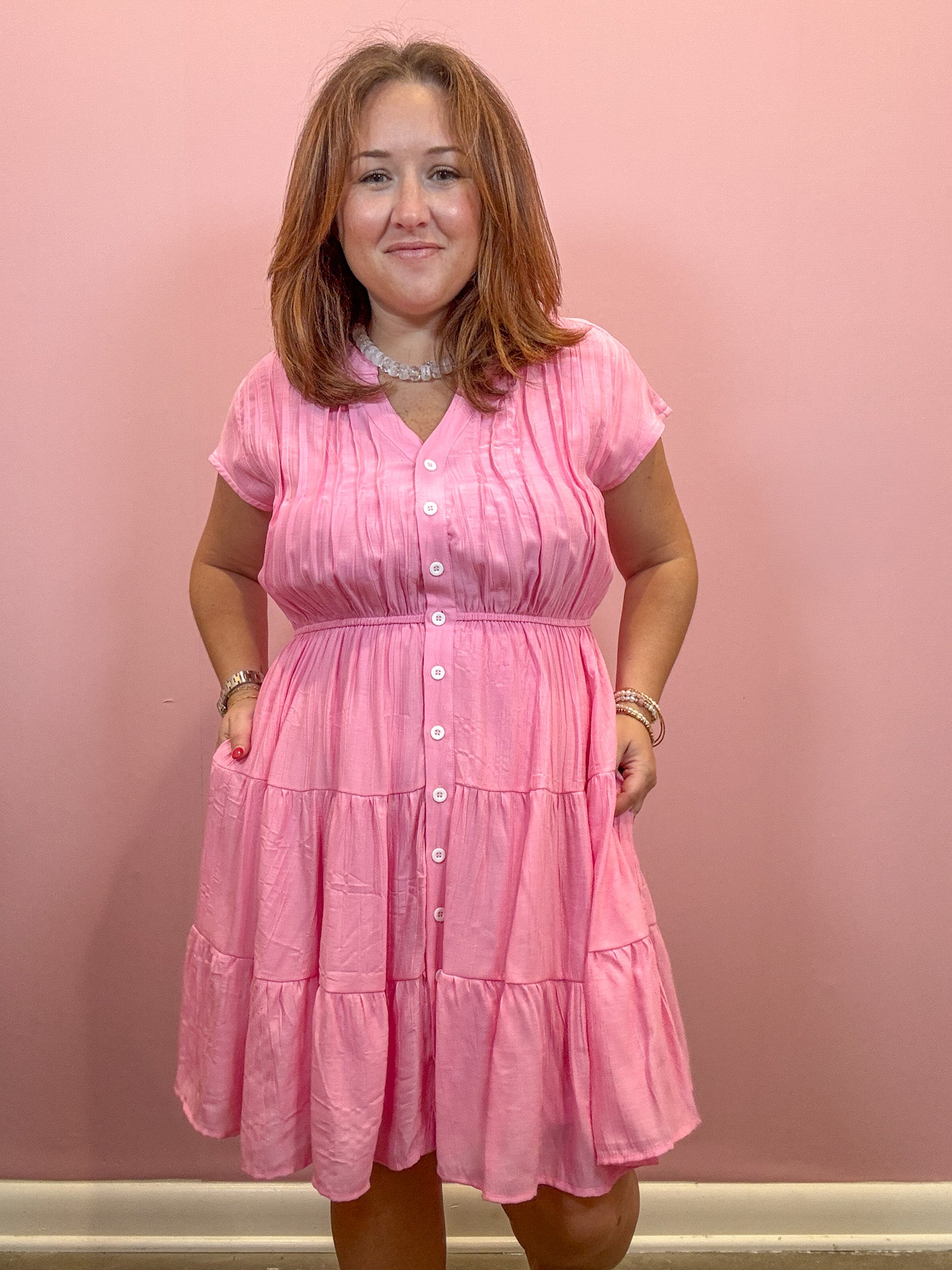 A woman wearing a pink tiered short-sleeve button-up dress, standing against a solid pink wall.