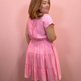 A woman wearing a pink tiered short-sleeve dress, photographed from the back against a solid pink wall.