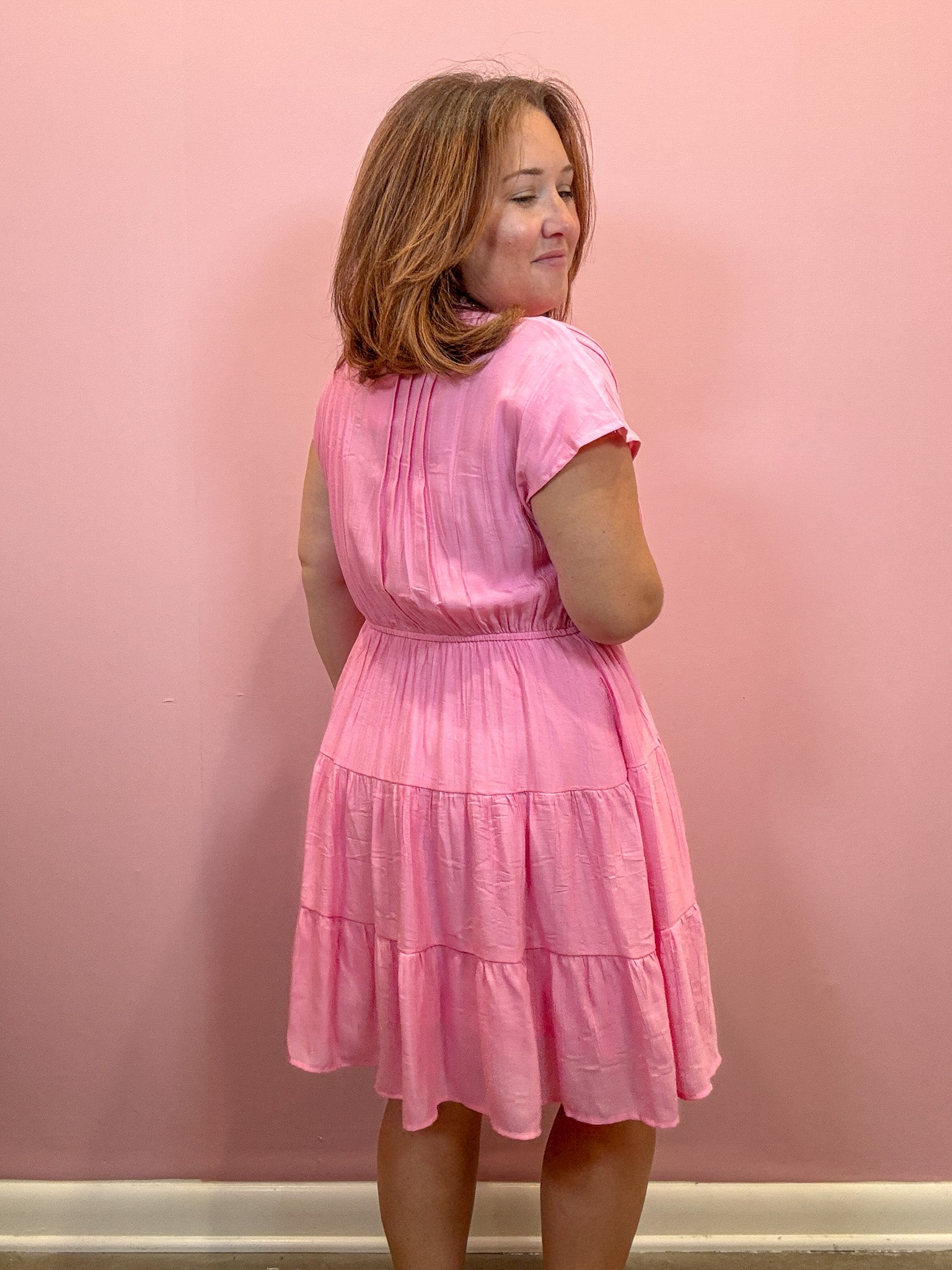 A woman wearing a pink tiered short-sleeve dress, photographed from the back against a solid pink wall.