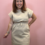 A woman modeling a light beige short-sleeve dress with a drawstring waist, standing against a solid pink wall.