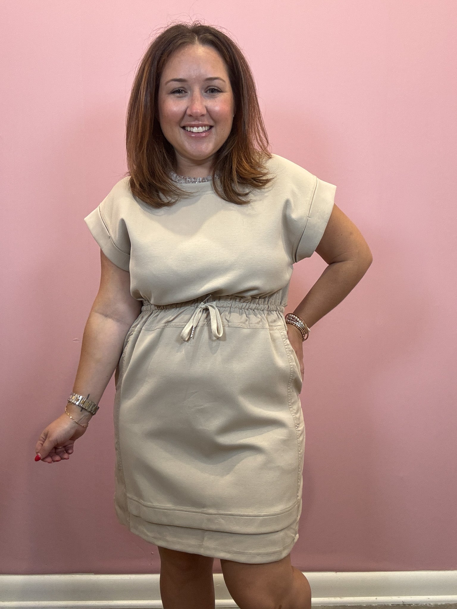A woman modeling a light beige short-sleeve dress with a drawstring waist, standing against a solid pink wall.