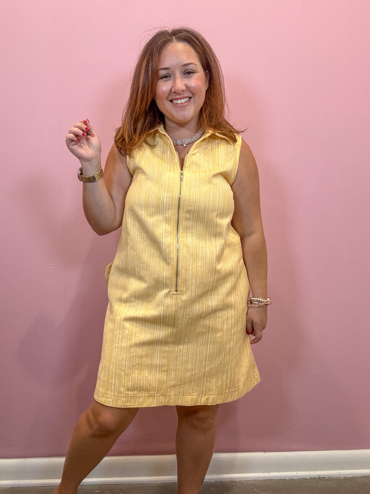 A woman modeling a sleeveless yellow pinstriped mini dress with a front zipper, standing against a solid pink wall.