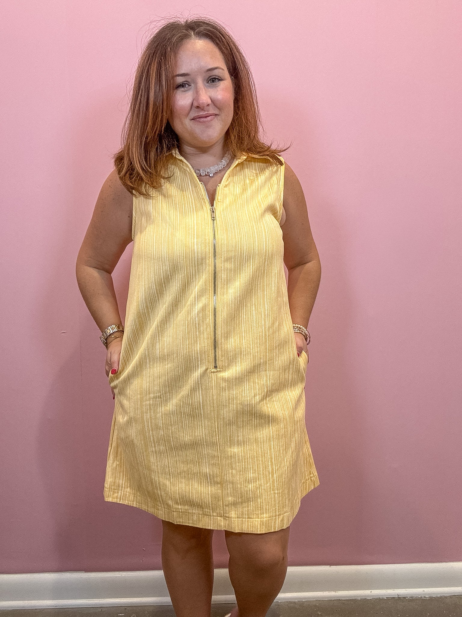 A woman modeling a sleeveless yellow textured dress with a front zipper and pockets, standing against a pink wall.