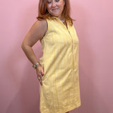 A woman modeling a sleeveless, yellow-and-white vertically striped mini dress with a zip-up front, standing against a solid pink wall.