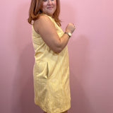 A woman wearing a sleeveless pale yellow dress with subtle vertical stripes, standing against a solid pink wall.