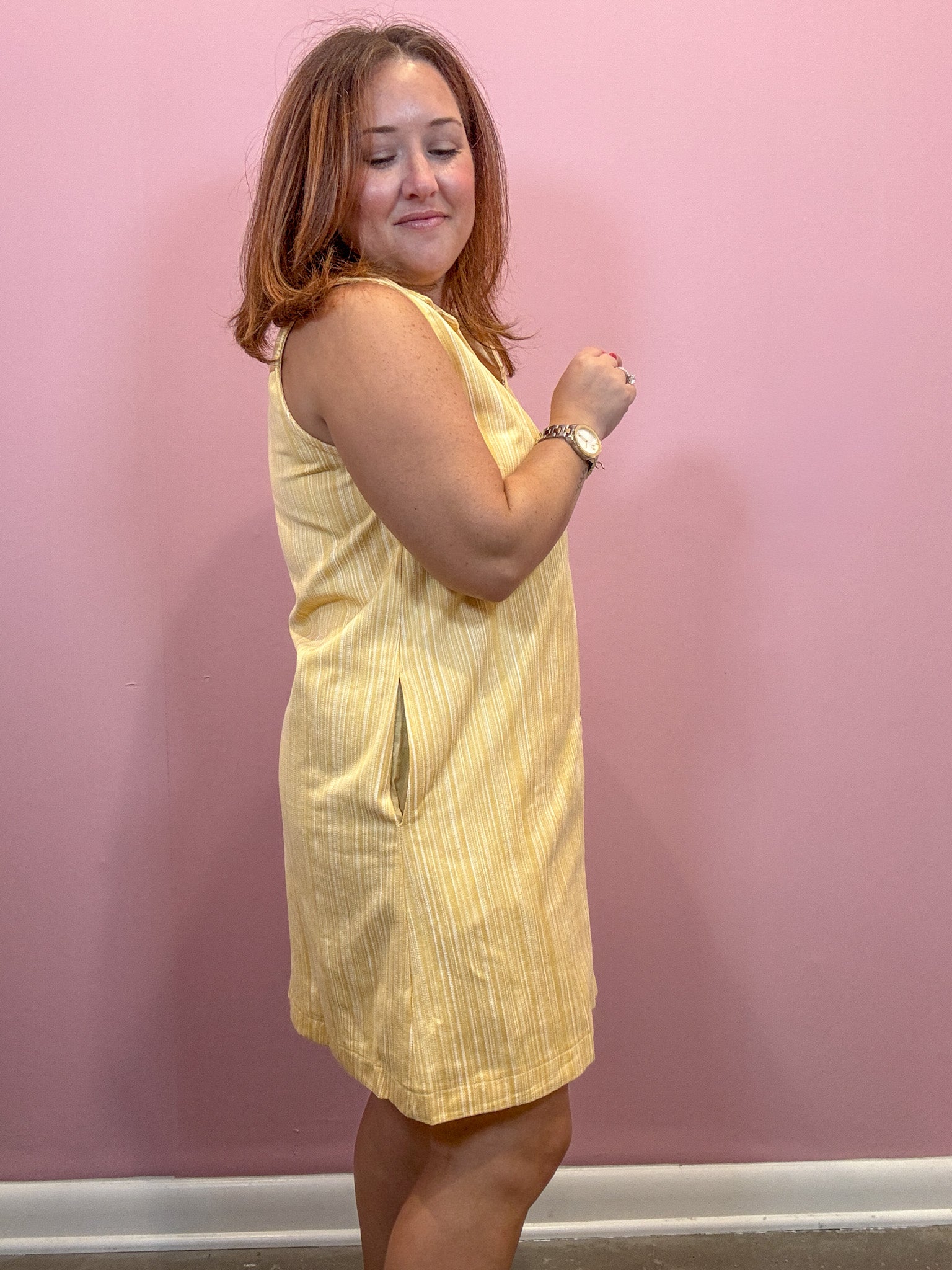 A woman wearing a sleeveless pale yellow dress with subtle vertical stripes, standing against a solid pink wall.