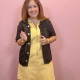 A woman modeling a yellow ribbed knit dress with a dark brown textured knit cardigan vest, standing against a solid pink wall.