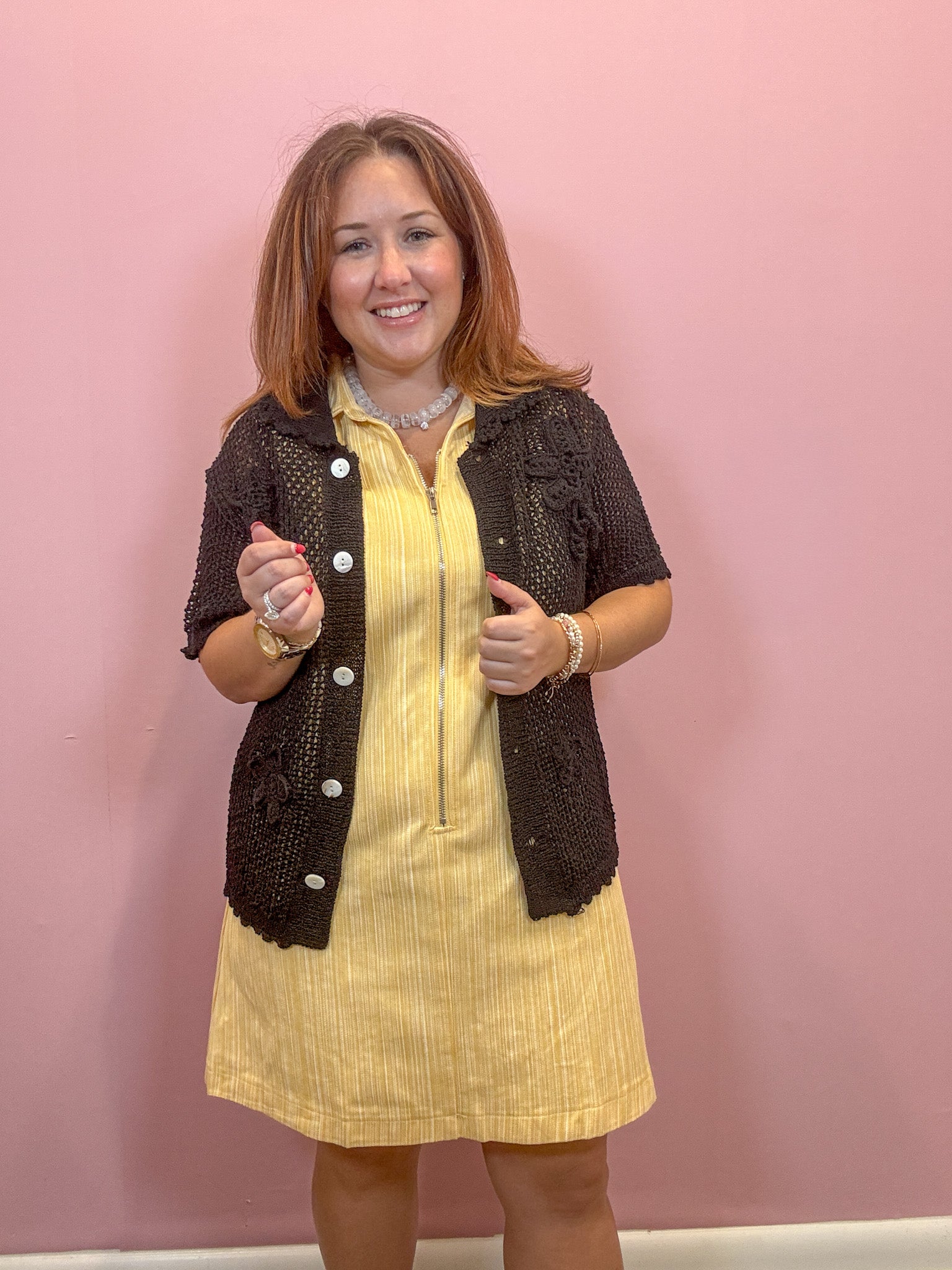 A woman modeling a yellow ribbed knit dress with a dark brown textured knit cardigan vest, standing against a solid pink wall.