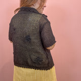 A woman shown from the back wearing a short-sleeve black crochet knit cardigan with textured floral appliqués and a scalloped hem, paired with a yellow pleated skirt, against a solid pink wall.