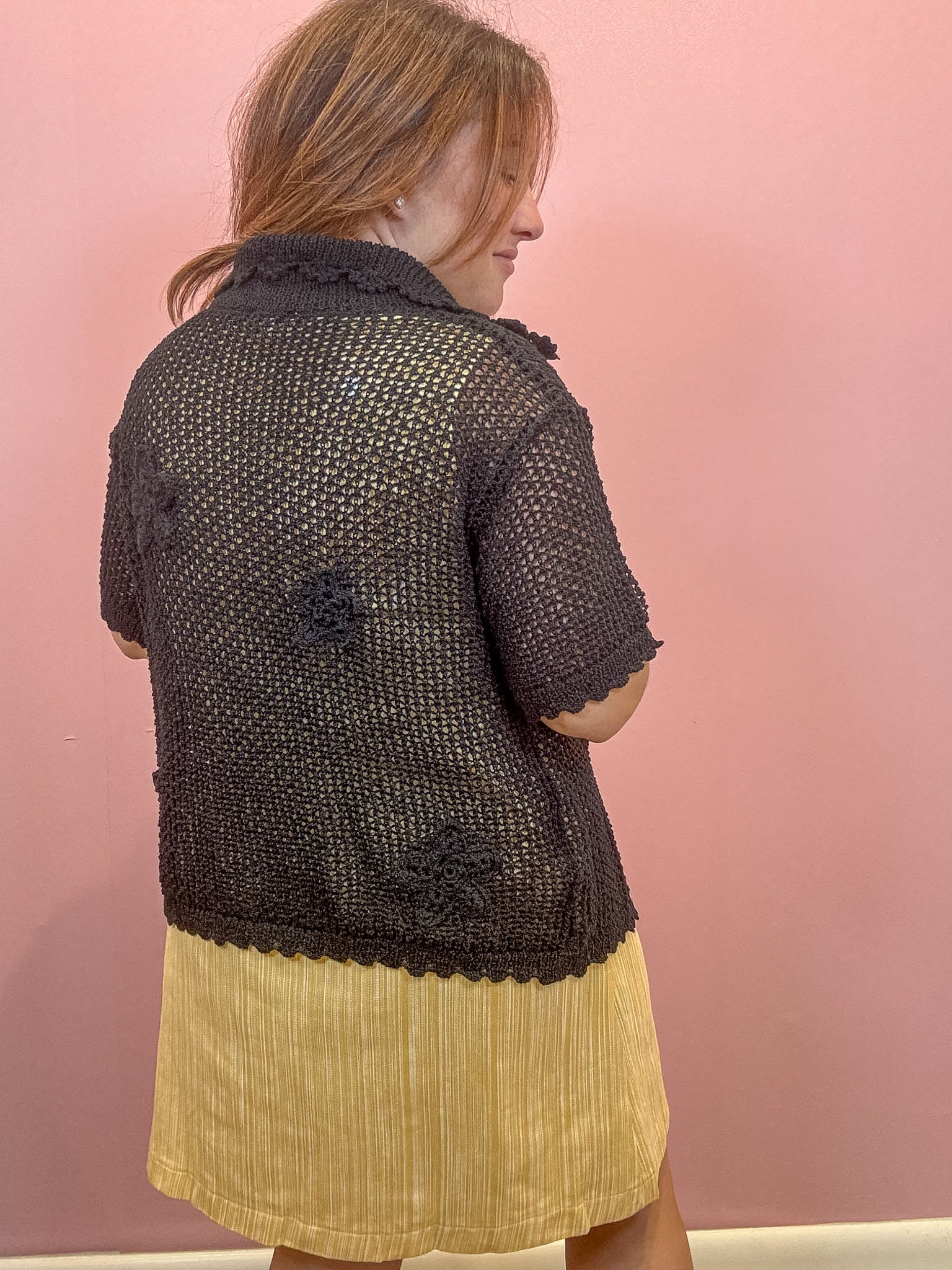 A woman shown from the back wearing a short-sleeve black crochet knit cardigan with textured floral appliqués and a scalloped hem, paired with a yellow pleated skirt, against a solid pink wall.