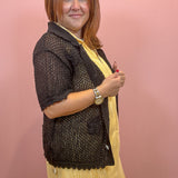 A woman wearing a black crochet knit cardigan over a yellow pleated dress, standing against a solid pink wall.