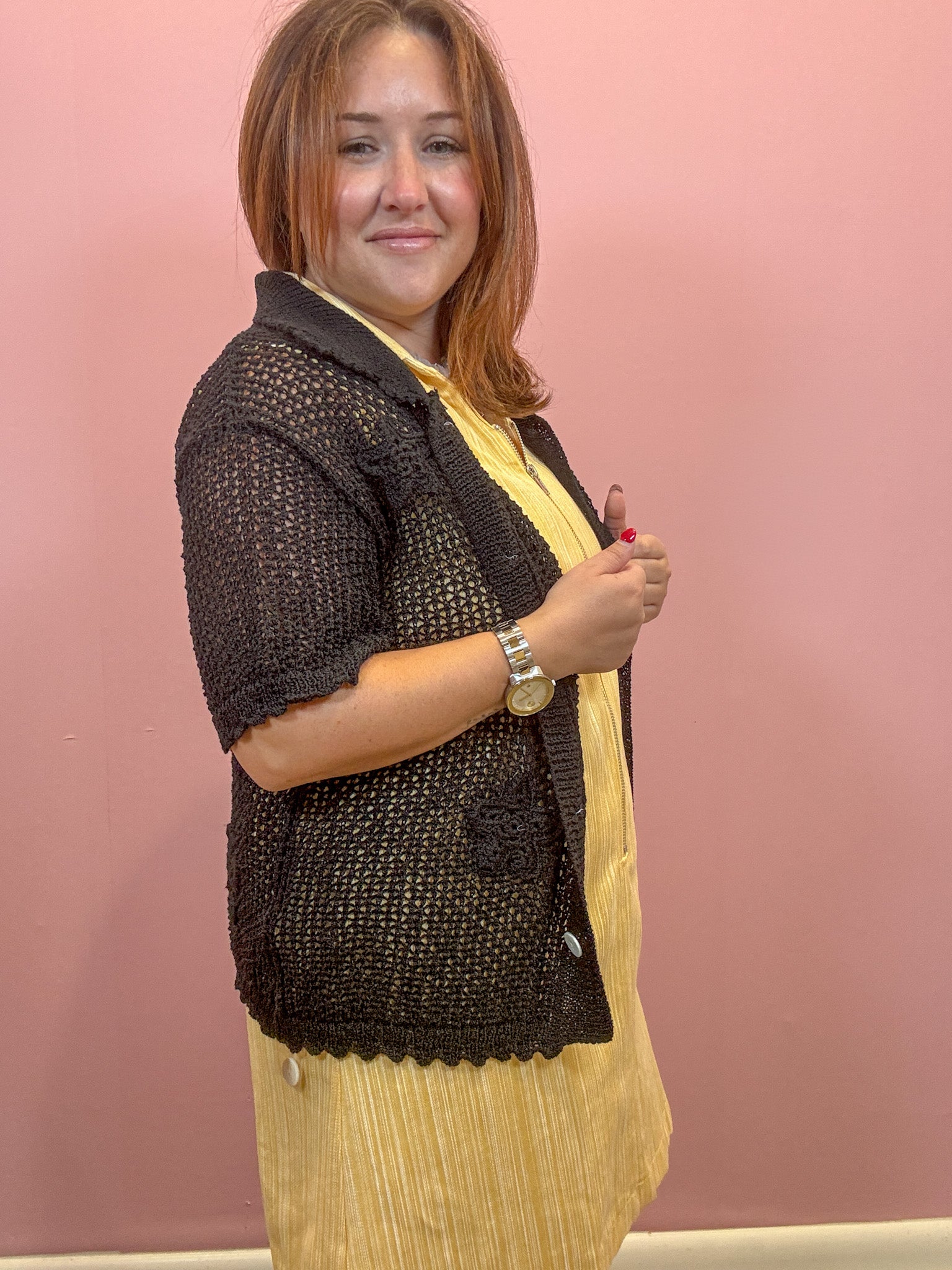 A woman wearing a black crochet knit cardigan over a yellow pleated dress, standing against a solid pink wall.
