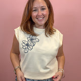 A smiling woman with shoulder-length brown hair stands against a solid pink background, wearing a cream-colored sleeveless t-shirt with a black floral line-art graphic on the left chest, paired with blue and white patterned pants.