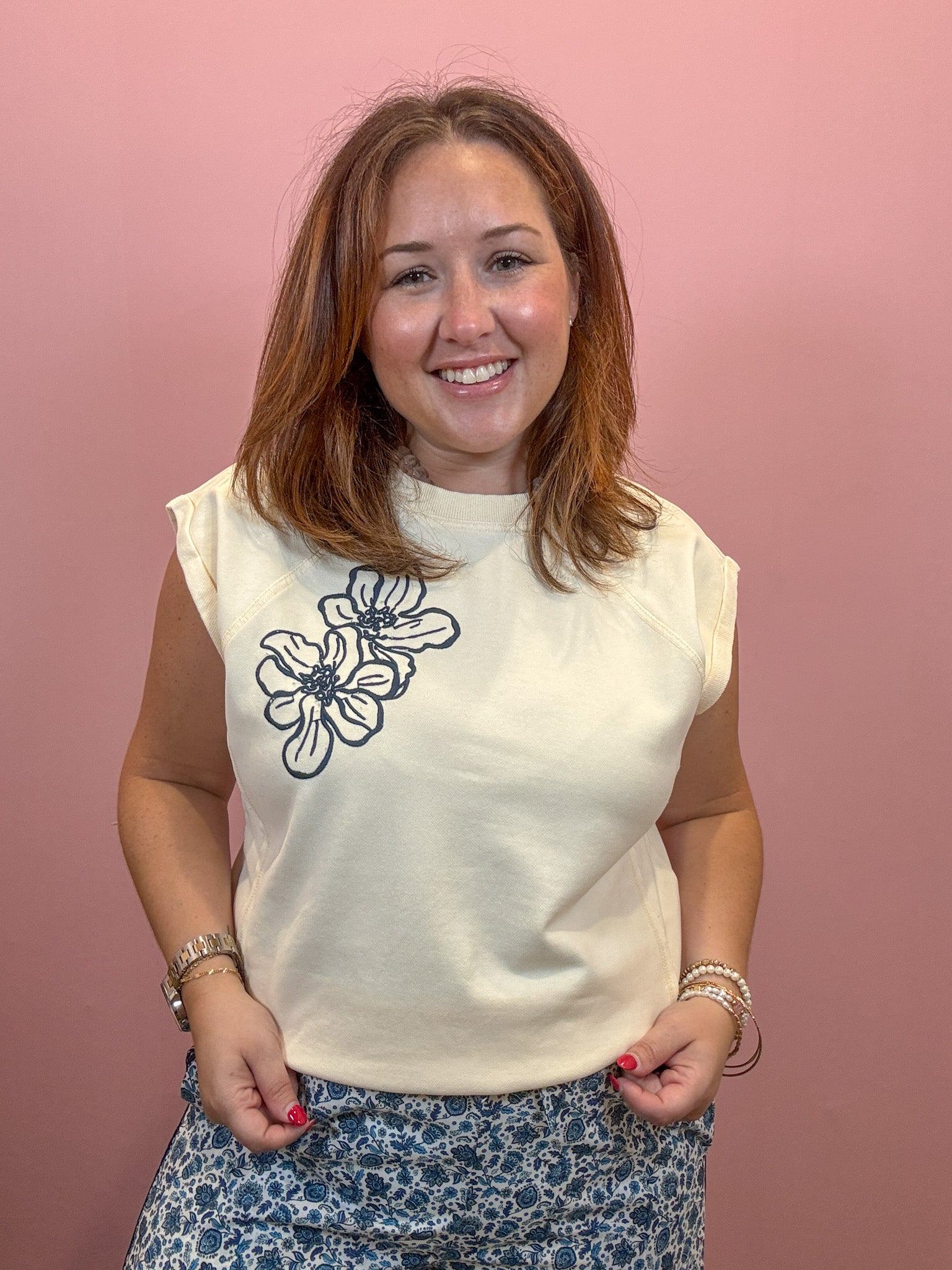 A smiling woman with shoulder-length brown hair stands against a solid pink background, wearing a cream-colored sleeveless t-shirt with a black floral line-art graphic on the left chest, paired with blue and white patterned pants.