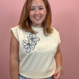 A smiling woman with shoulder-length auburn hair stands against a solid pink wall, wearing a sleeveless cream-colored top with a black floral line-art graphic on the left chest, paired with blue and white patterned bottoms.