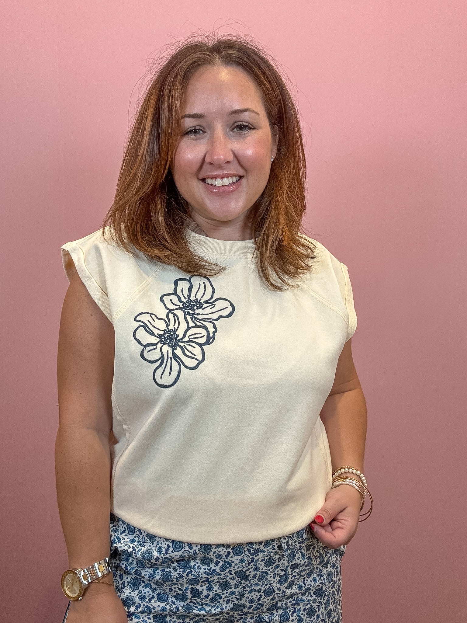 A smiling woman with shoulder-length auburn hair stands against a solid pink wall, wearing a sleeveless cream-colored top with a black floral line-art graphic on the left chest, paired with blue and white patterned bottoms.