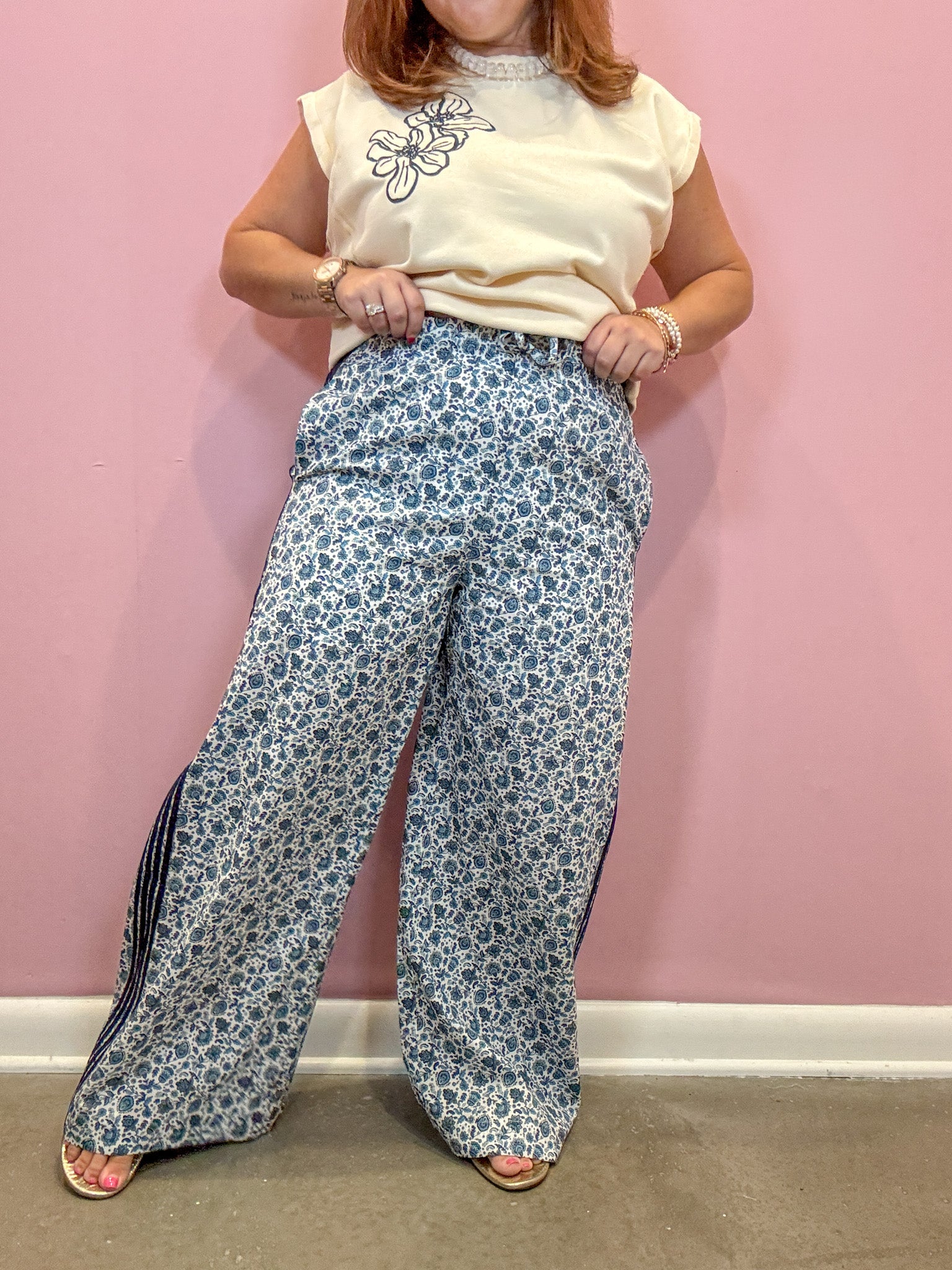 A person modeling wide-leg pants with a blue and white floral pattern and side slits, paired with a cream sleeveless top, standing against a pink wall.