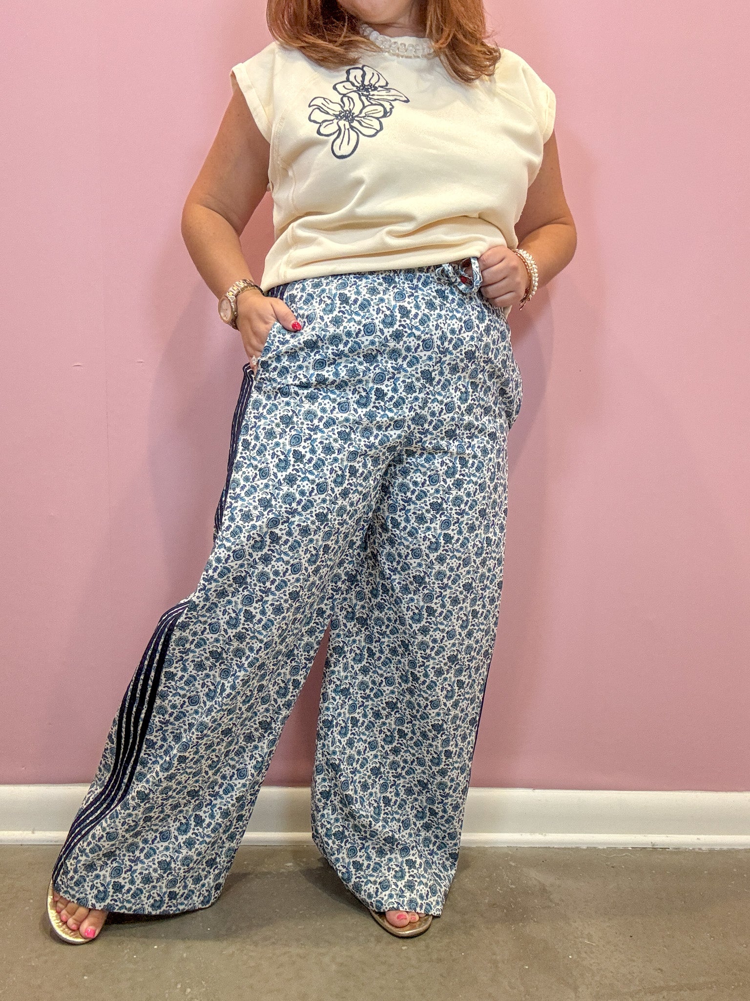 A person modeling wide-leg pants with a blue and white floral print and dark side stripes, paired with a cream sleeveless top, standing against a pink wall.
