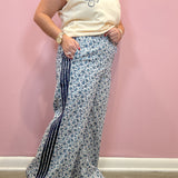 A woman modeling a pair of wide-leg, high-waisted pants featuring a blue and white floral pattern with dark navy side stripes, paired with a cream sleeveless top against a pink wall.