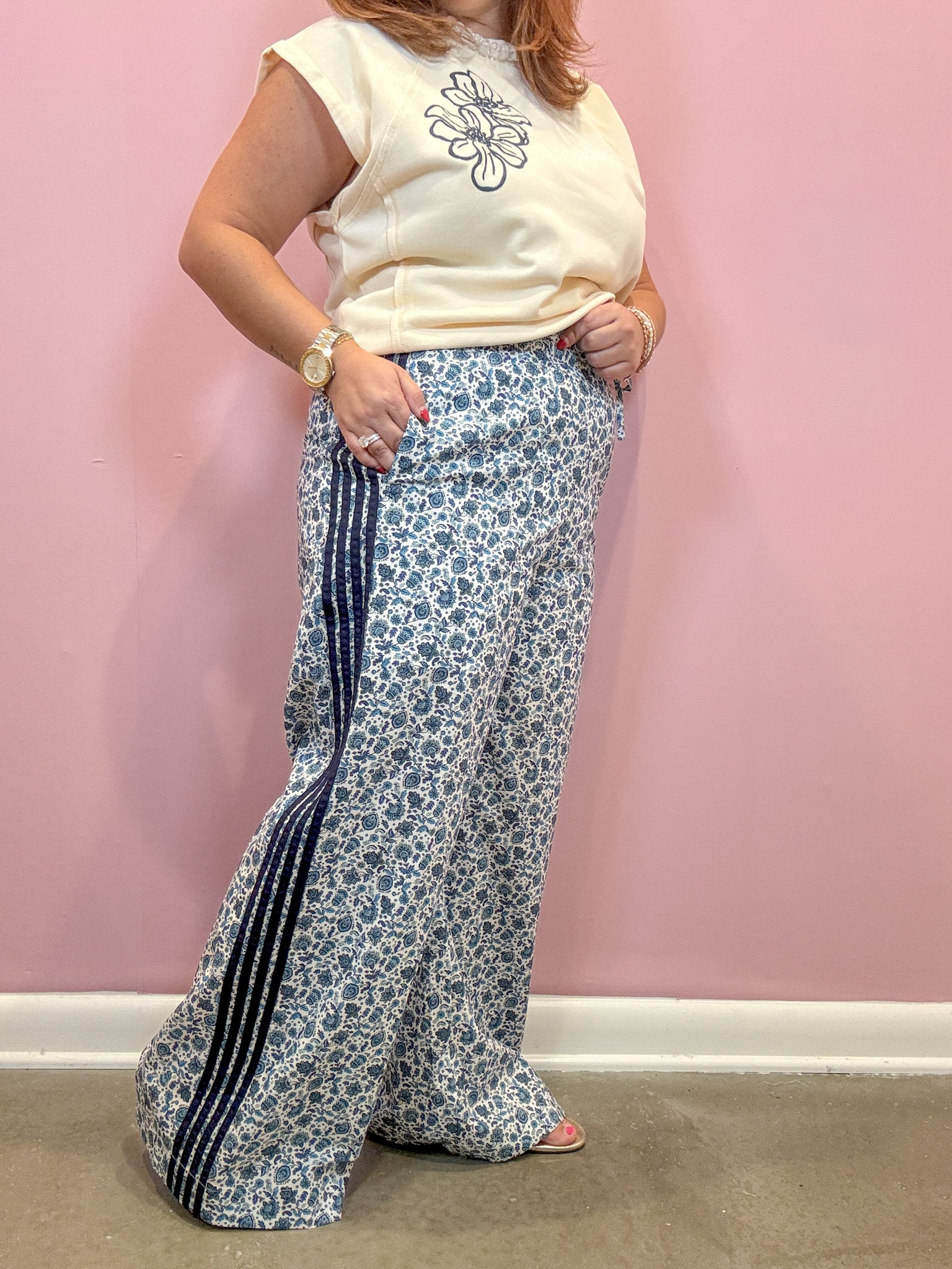 A woman modeling a pair of wide-leg, high-waisted pants featuring a blue and white floral pattern with dark navy side stripes, paired with a cream sleeveless top against a pink wall.