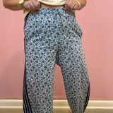 A person wearing loose, wide-leg pants with a blue and white floral print and dark navy side stripes, paired with a cream t-shirt, standing against a pink wall.