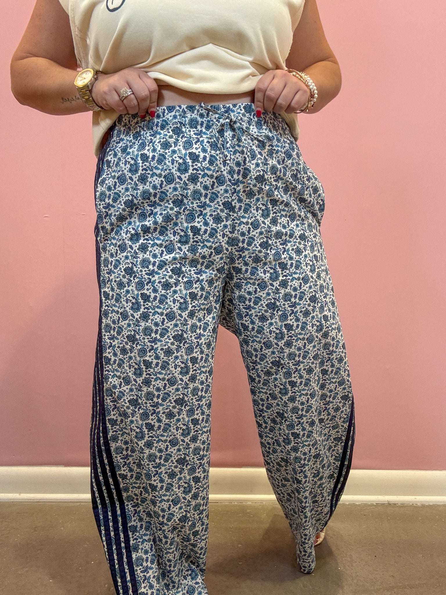 A person wearing loose, wide-leg pants with a blue and white floral print and dark navy side stripes, paired with a cream t-shirt, standing against a pink wall.