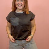 A woman with light brown hair smiling, wearing a dark brown short-sleeve t-shirt with a textured gray chest pocket and gray pinstriped trousers, standing against a solid pink wall.