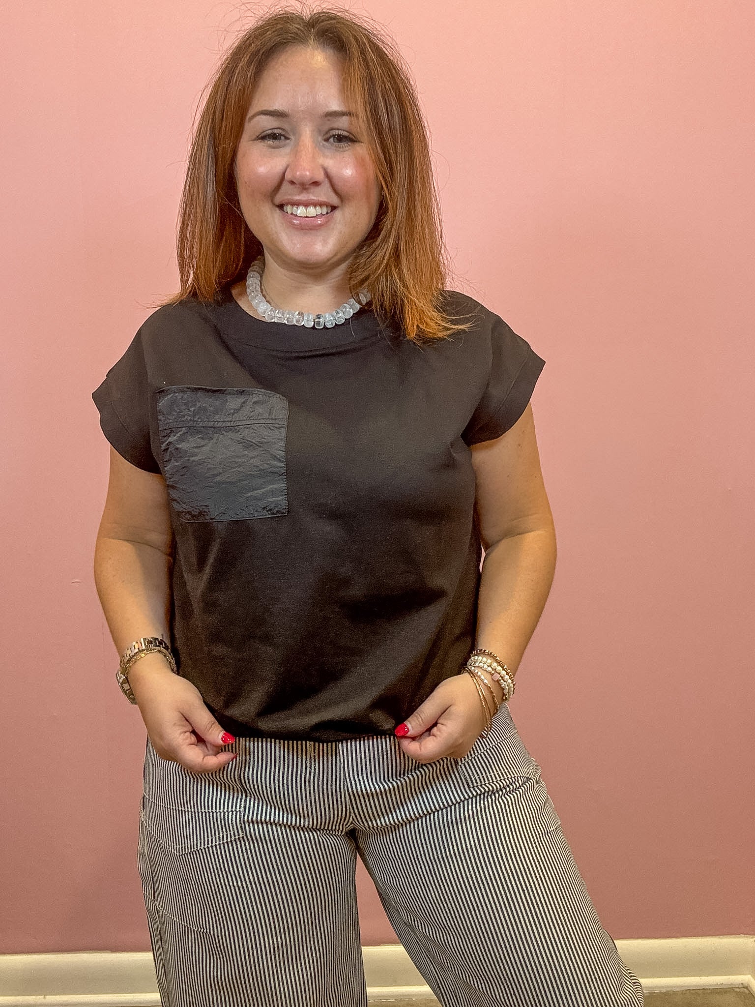 A woman with light brown hair smiling, wearing a dark brown short-sleeve t-shirt with a textured gray chest pocket and gray pinstriped trousers, standing against a solid pink wall.