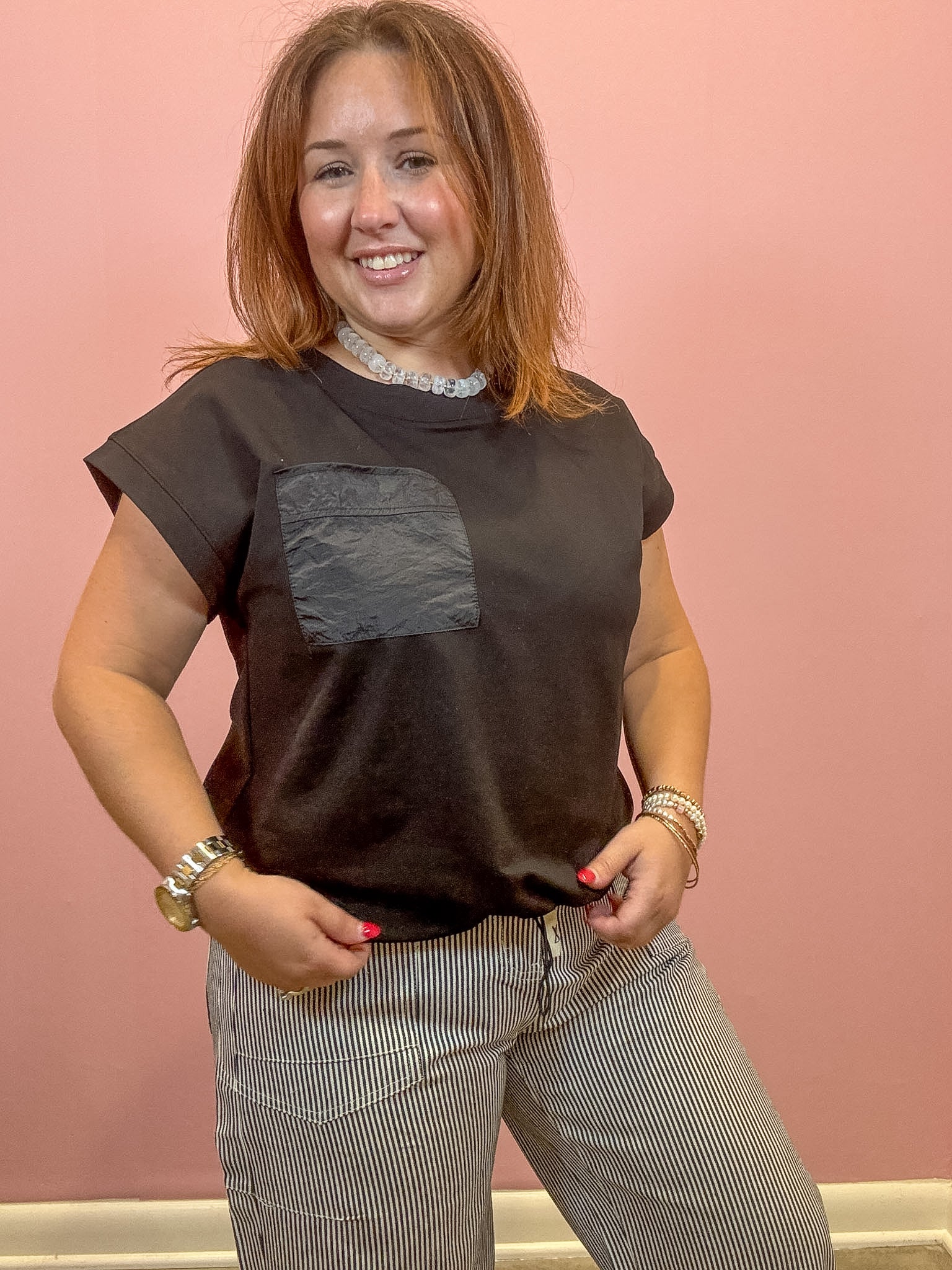 A woman modeling a dark brown short-sleeve t-shirt with a textured gray chest pocket, paired with gray and white pinstriped trousers, standing against a solid pink wall.