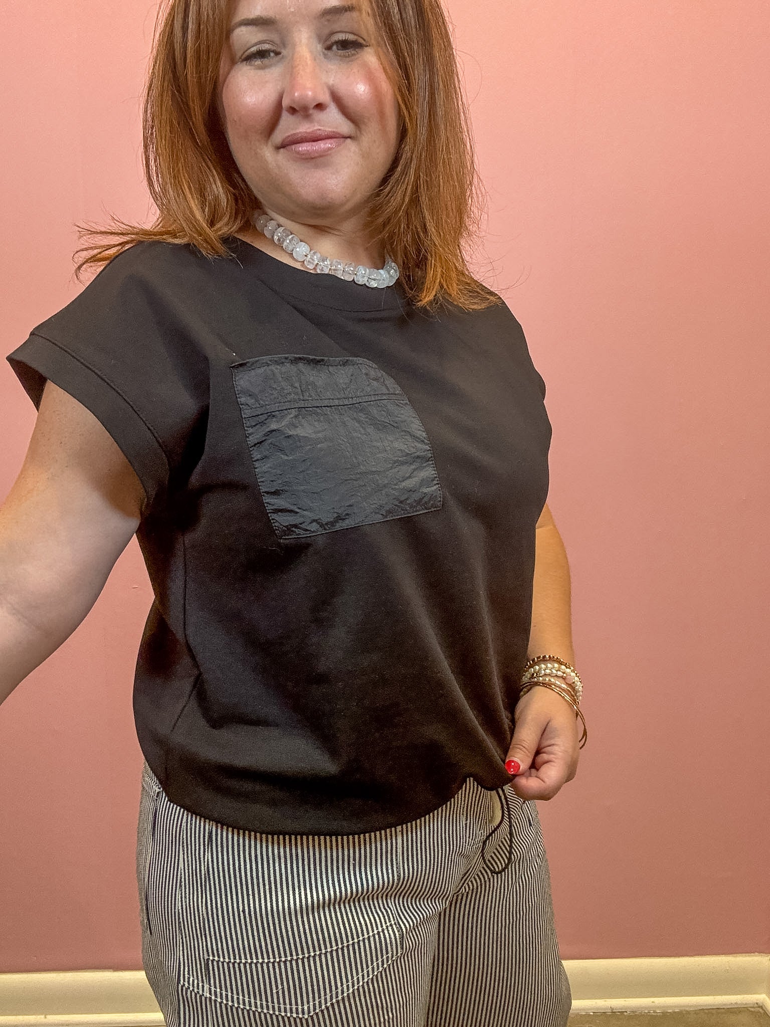 A woman wearing a dark brown short-sleeve t-shirt with a contrasting dark gray chest pocket, paired with gray and white striped pants, standing against a solid pink wall.