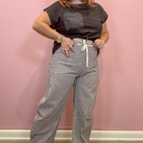 A woman modeling a pair of gray and white pinstriped wide-leg pants with a cream drawstring, paired with a dark brown t-shirt, standing against a pink wall.
