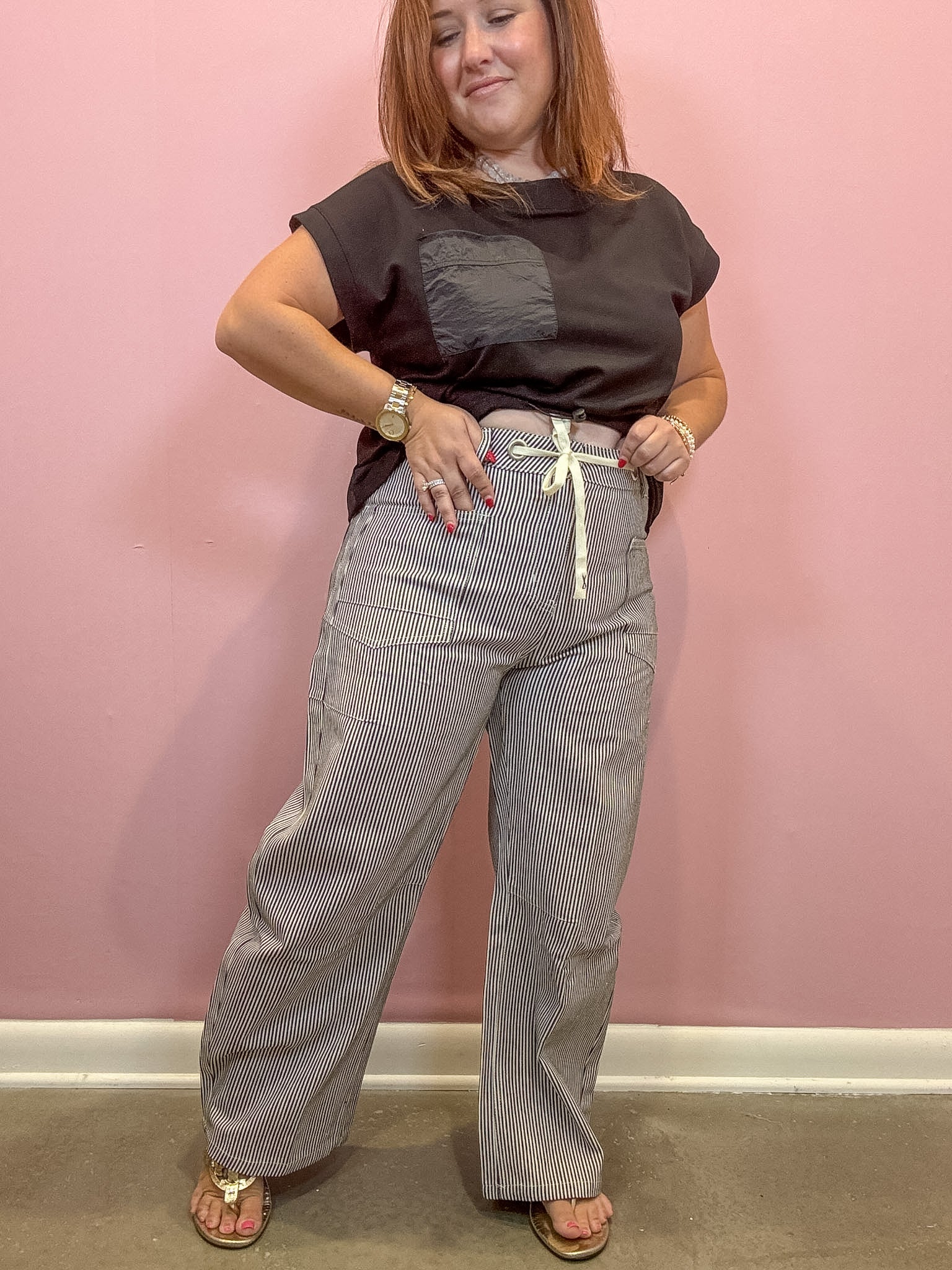 A woman modeling a pair of gray and white pinstriped wide-leg pants with a cream drawstring, paired with a dark brown t-shirt, standing against a pink wall.
