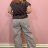 A person viewed from behind wearing a dark charcoal gray t-shirt and light gray and white pinstriped wide-leg pants, standing against a solid pink wall.
