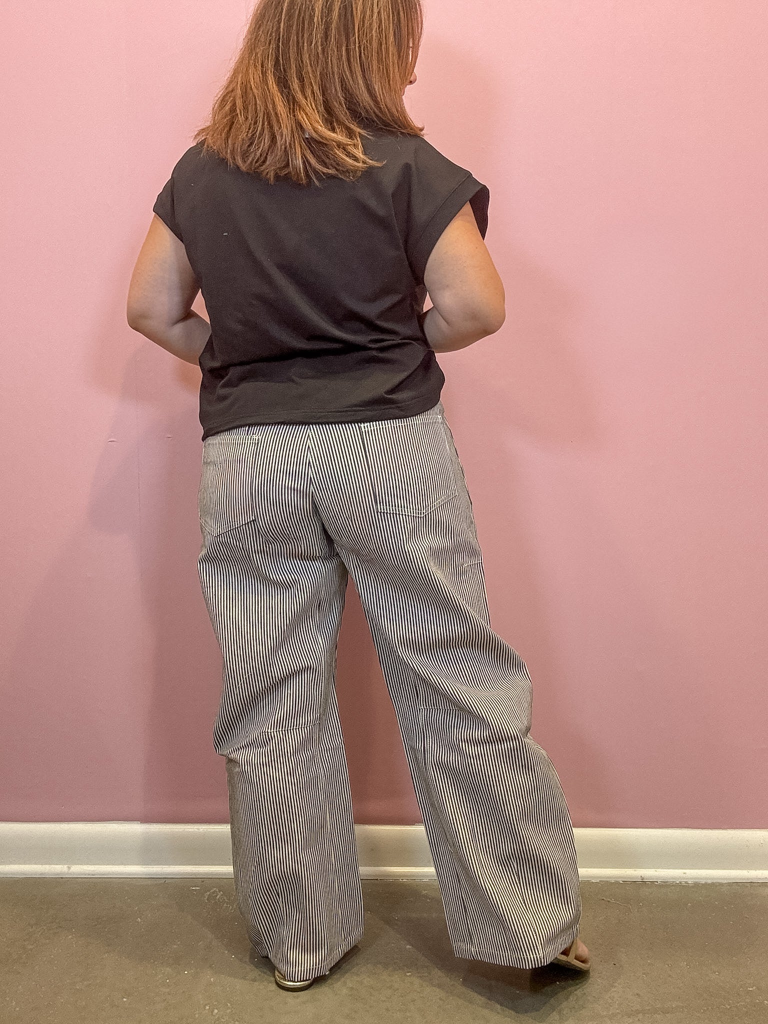 A person viewed from behind wearing a dark charcoal gray t-shirt and light gray and white pinstriped wide-leg pants, standing against a solid pink wall.