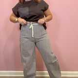 A woman modeling a pair of wide-leg gray pinstriped pants with a white drawstring, paired with a dark brown t-shirt, standing against a pink wall.