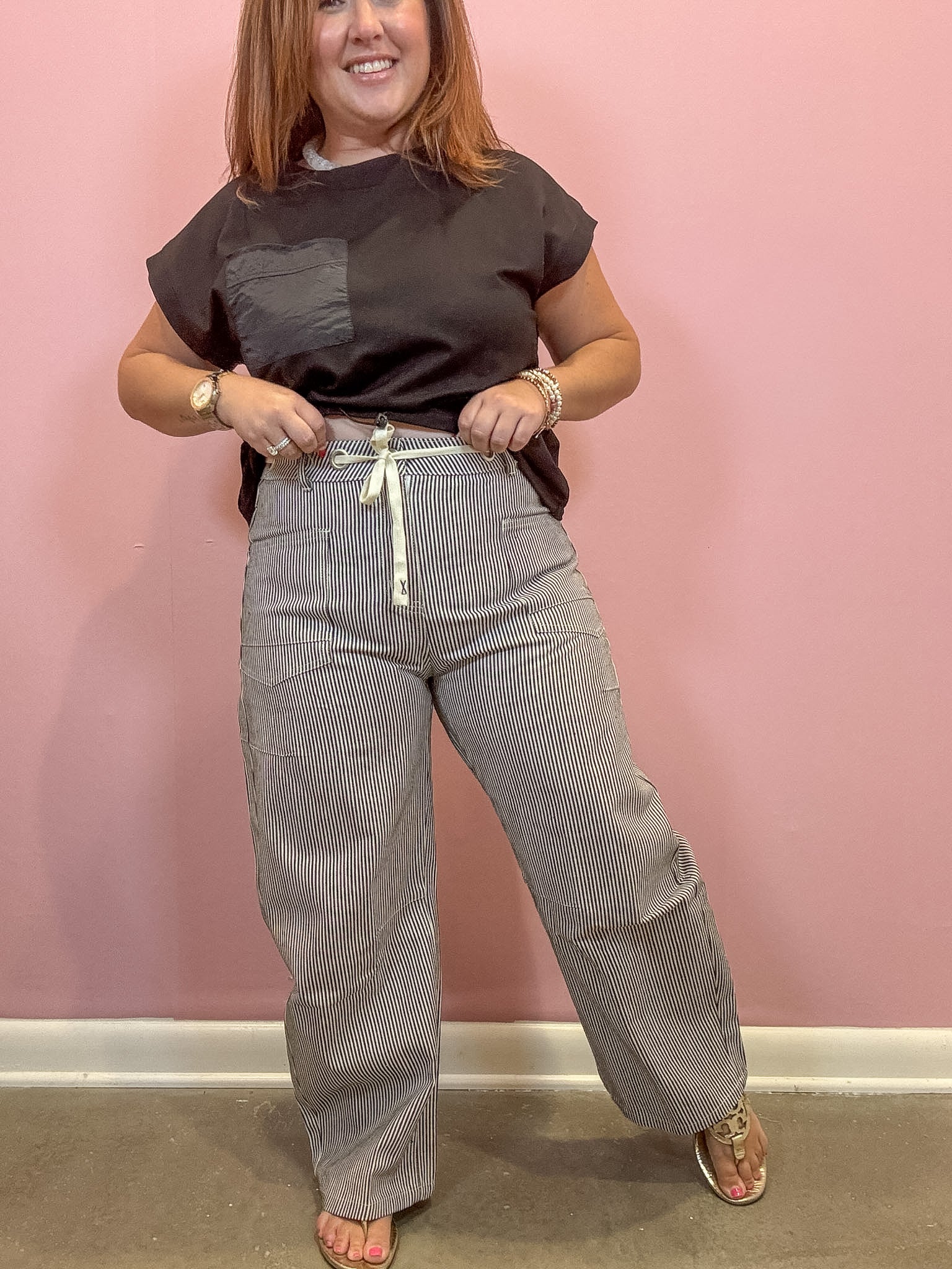 A woman modeling a pair of wide-leg gray pinstriped pants with a white drawstring, paired with a dark brown t-shirt, standing against a pink wall.