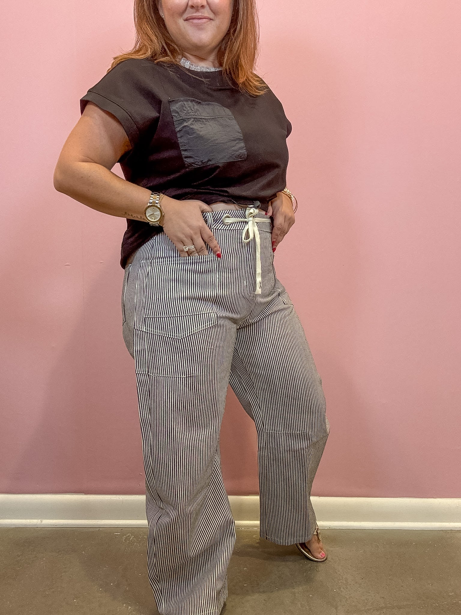 A woman modeling a dark brown short-sleeve t-shirt with a chest pocket and high-waisted, wide-leg gray pinstriped pants with a cream drawstring, standing against a pink wall.