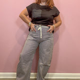 A woman modeling a dark brown short-sleeve t-shirt with a chest pocket and gray pinstriped wide-leg pants with a white drawstring, standing against a pink wall.