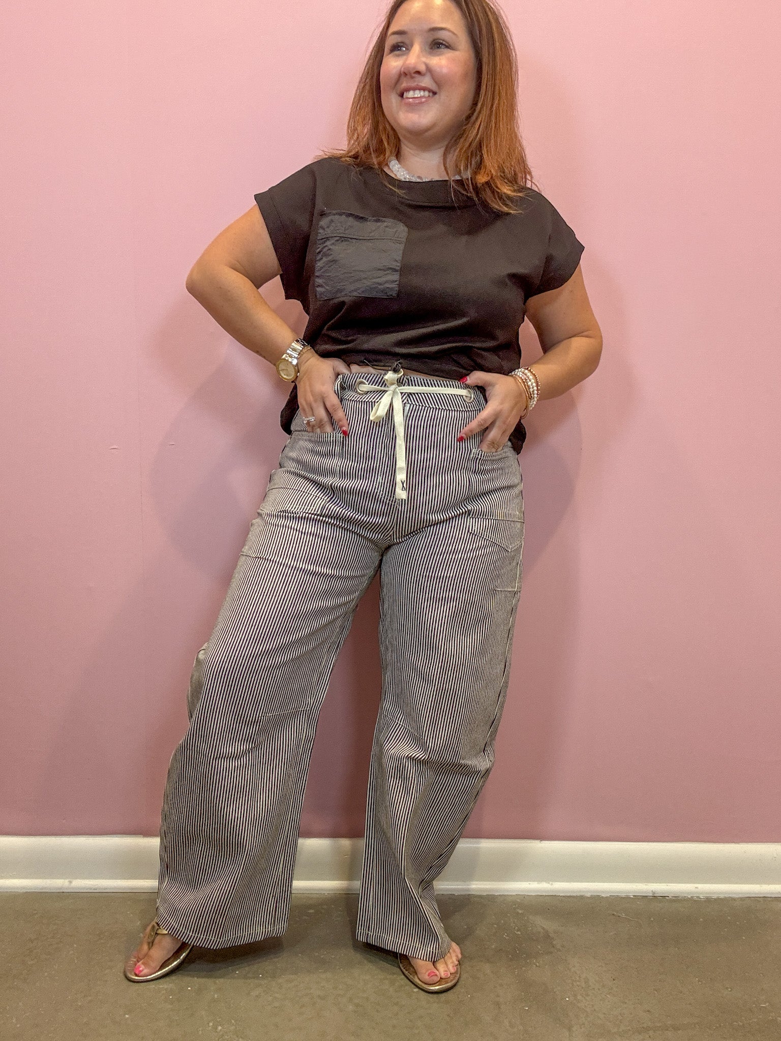 A woman modeling a dark brown short-sleeve t-shirt with a chest pocket and gray pinstriped wide-leg pants with a white drawstring, standing against a pink wall.