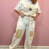 A woman modeling a white short-sleeve top with a large beige flower applique on the front and matching white drawstring pants with beige patch pockets, standing against a pink wall.