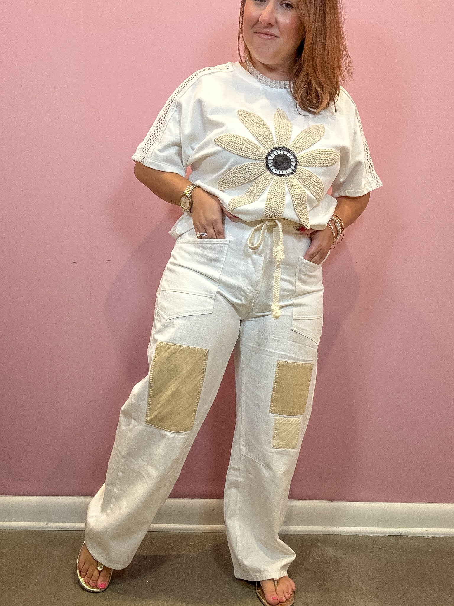 A woman modeling a white short-sleeve top with a large beige flower applique on the front and matching white drawstring pants with beige patch pockets, standing against a pink wall.