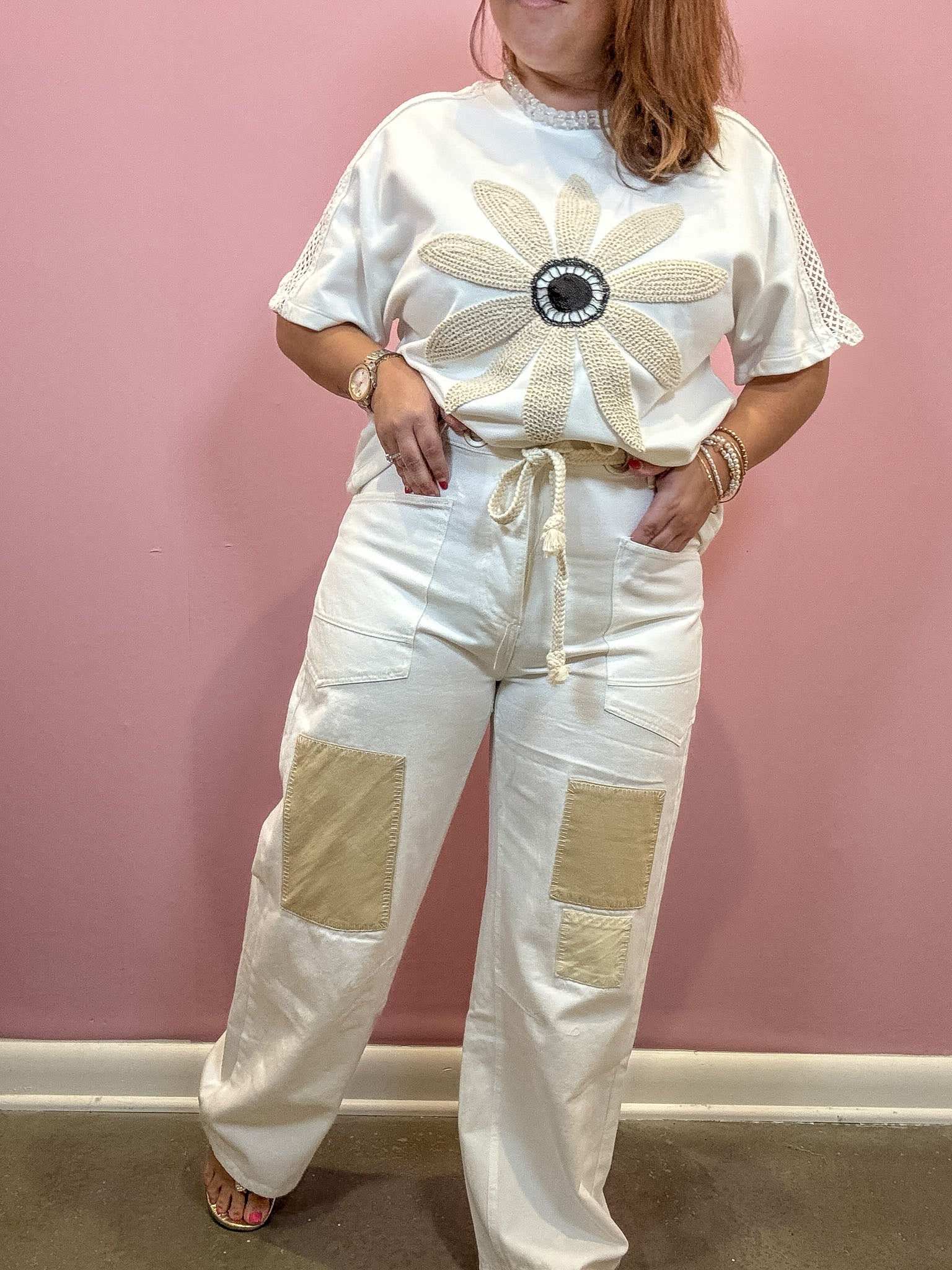 A woman wearing a white short-sleeve top with a large beige flower applique on the chest and matching white wide-leg pants with beige patch pockets, standing against a pink wall.