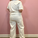 A woman wearing a white short-sleeve top and matching wide-leg pants, shown from the back against a pink wall.
