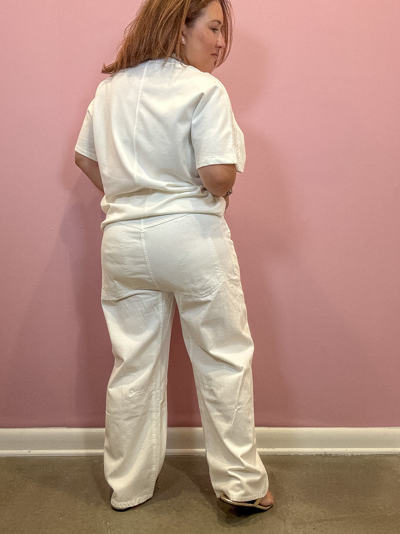 A woman wearing a white short-sleeve top and matching wide-leg pants, shown from the back against a pink wall.