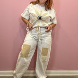 A woman modeling a white short-sleeve t-shirt and matching wide-leg pants set against a pink wall. The outfit features a large beige flower applique on the chest with a black and white center, beige drawstrings at the waist, and two large beige patch pockets on the pants.