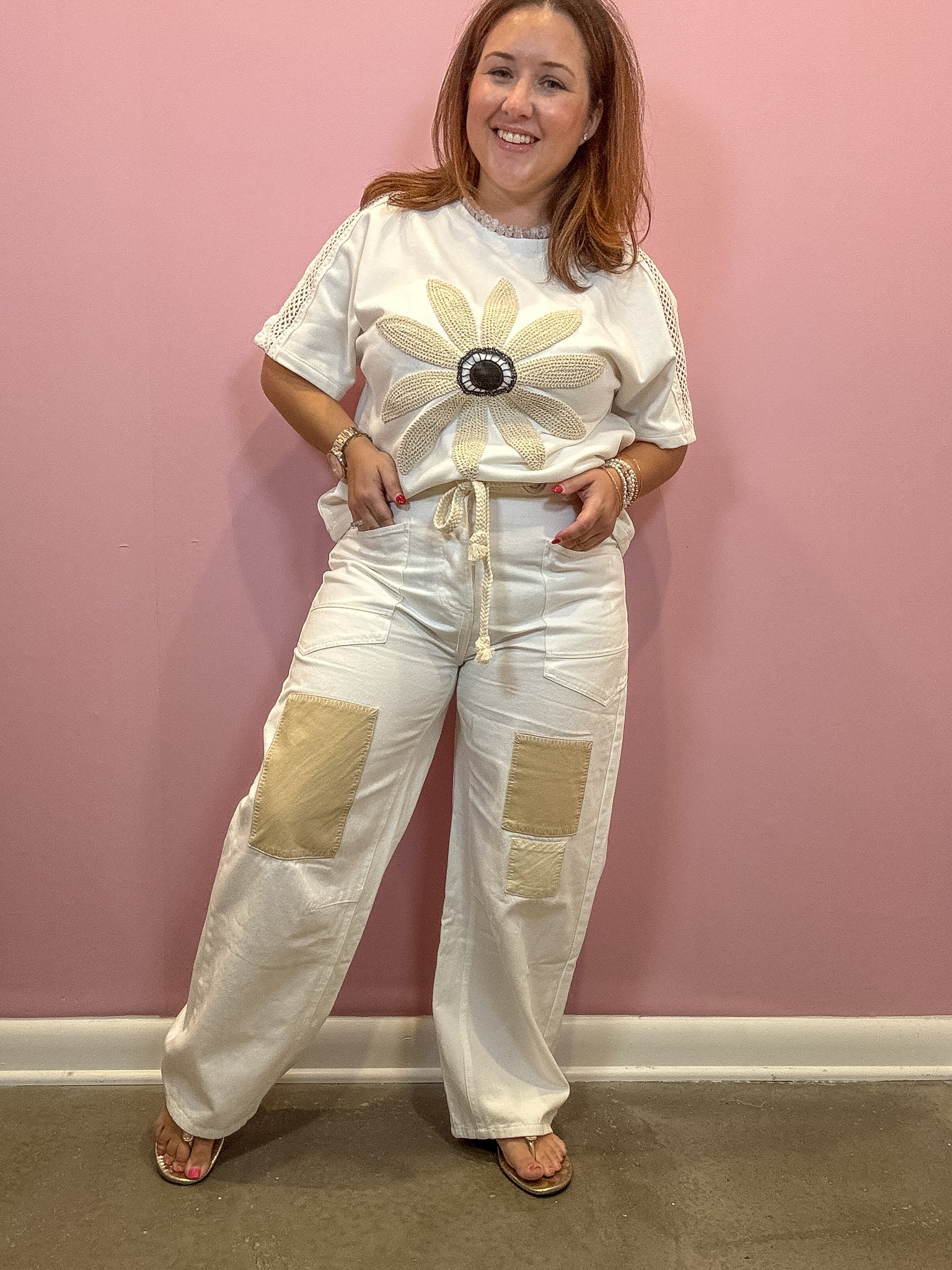 A woman modeling a white short-sleeve t-shirt and matching wide-leg pants set against a pink wall. The outfit features a large beige flower applique on the chest with a black and white center, beige drawstrings at the waist, and two large beige patch pockets on the pants.