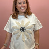 A woman wearing a white short-sleeve t-shirt with a large beige beaded flower design on the front, paired with beige pants, standing against a solid pink wall.