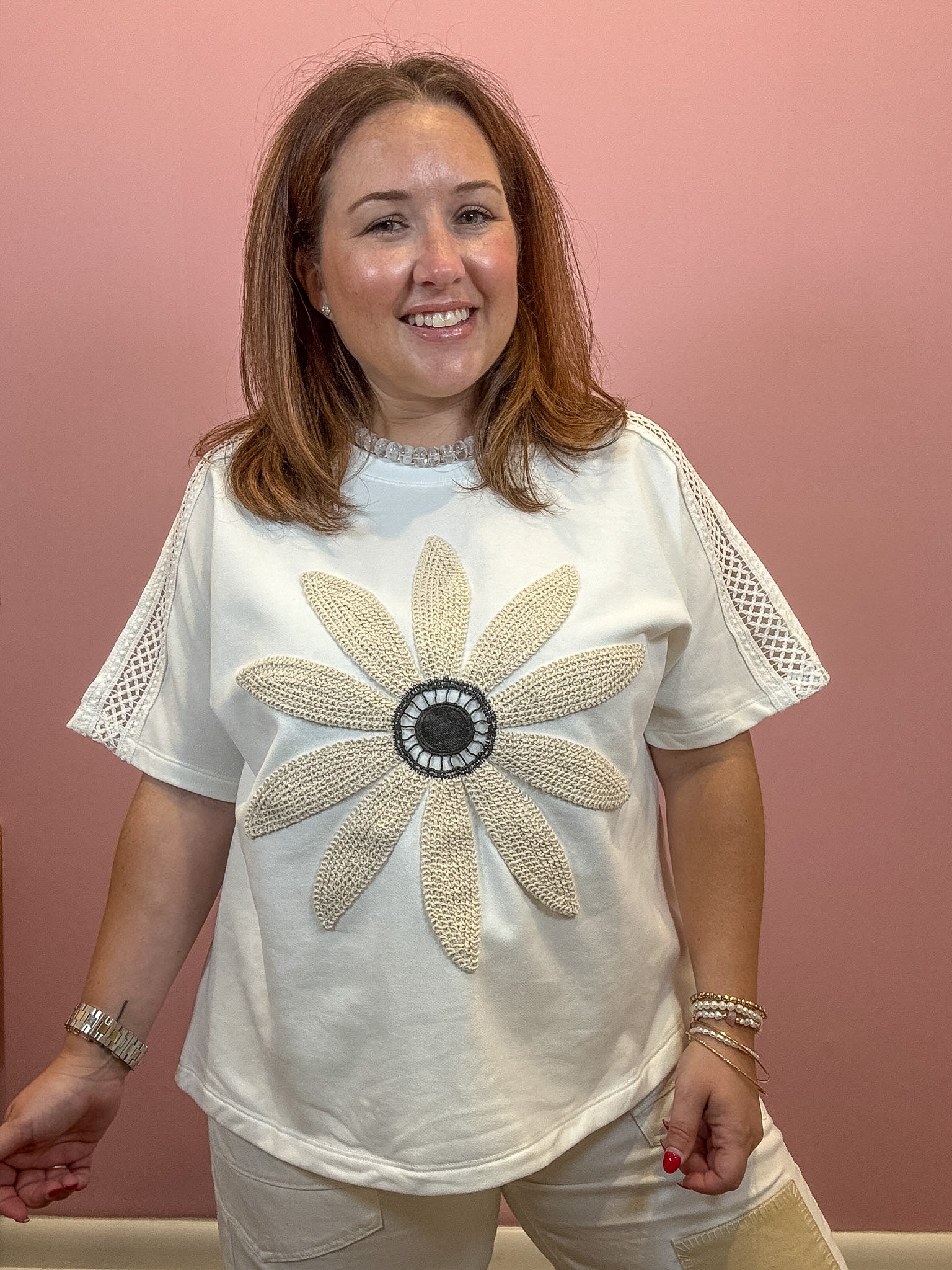 A woman wearing a white short-sleeve t-shirt with a large beige beaded flower design on the front, paired with beige pants, standing against a solid pink wall.